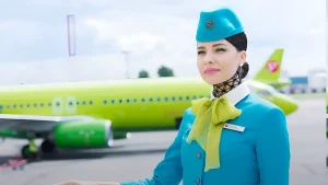 Стюардесса S7 Airlines в бирюзовой форме с салатовым шейным платком стоит на фоне самолета.