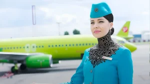 Стюардесса S7 Airlines в форме стоит на фоне самолета с ярким салатовым фюзеляжем. На шее стюардессы видна сложная геометрическая татуировка в стиле blackwork.