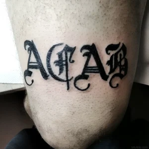 Татуировка аббревиатуры ACAB, выполненная черным цветом в готическом стиле на коже человека.
