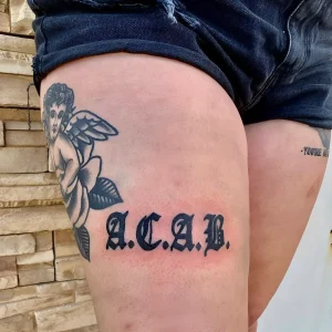 Фото тату ACAB, выполненное в готическом стиле, на бедре человека, рядом с изображением ангела и розы.