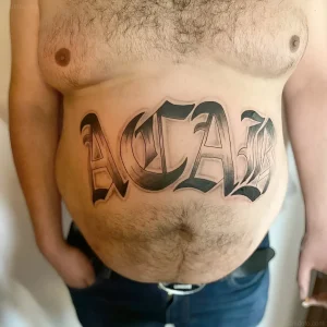 Крупное тату "ACAB" в готическом стиле, выполненное черными чернилами, расположено горизонтально на животе мужчины.