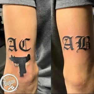 Татуировка "AC" с силуэтом пистолета на одном предплечье и "AB" готическим шрифтом на другом.
