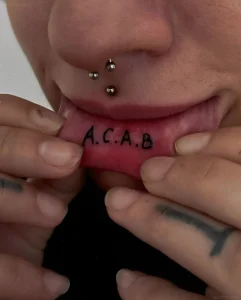 Макрофото татуировки "A.C.A.B." на внутренней стороне нижней губы, демонстрирующее четкие черные буквы на розовой слизистой.