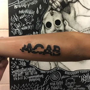 Крупный план татуировки "ACAB" на предплечье, выполненной в черном цвете, с элементами протестной графики на фоне.