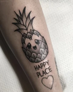Татуировка милого ананаса с большими глазами и улыбкой, рядом надпись 'Happy Place' и контур сердца