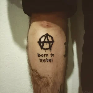 Татуировка символа анархии с надписью "Born to Rebel" на ноге