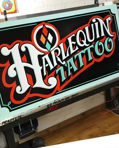 Яркая вывеска с надписью "Harlequin Tattoo" в стиле старой школы, выполненная в контрастных цветах