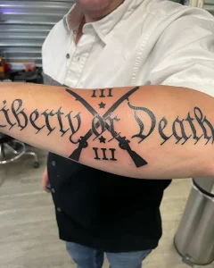 Татуировка "Liberty or Death" с двумя перекрещенными мушкетами и римскими цифрами III, выполненная черными чернилами на предплечье
