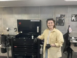 Grace DeVrieze, owner of Alchemist Art Studio in downtown Rock Island, stands next to black tattoo equipment. She is wearing a yellow shirt and black gloves, smiling at the camera.