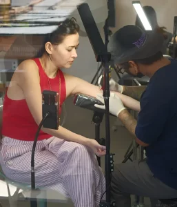 Актриса Бела Падила сидит в студии Tattoo Nebula, пока мастер наносит ей на предплечье тонкую татуировку белым пигментом. На фото видны профессиональное освещение и процесс работы над деликатным узором.