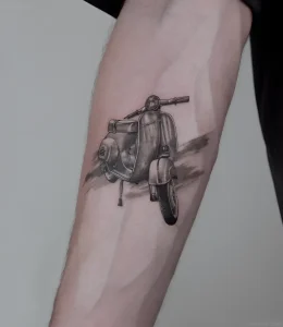 Детальная черно-белая татуировка ретро-скутера Vespa с эффектом движения на мужском предплечье, выполненная в стиле реализма с акцентом на текстуры и динамику