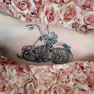 Детальная черно-белая татуировка классического мотоцикла Harley-Davidson на предплечье человека, выполненная в стиле American Traditional с четкими контурами и штриховкой, на фоне розовых роз