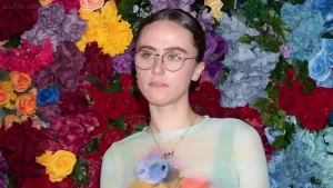Портрет Эммы Эмхофф в очках и пастельной блузе на фоне яркой стены из цветов