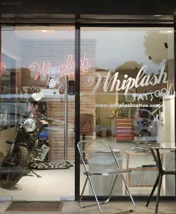Фотография входа в тату-студию Whiplash Tattoo через стеклянные двери. Внутри виден мотоцикл, неоновая вывеска и мебель.