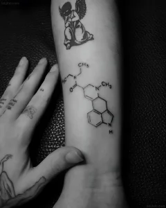 Черно-белое фото предплечья с татуировкой химической формулы MDMA в стиле лайнворк, рядом видна татуировка ангела и рука с надписями