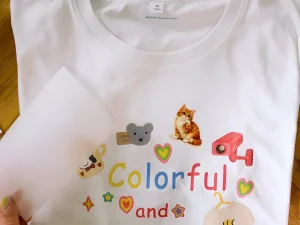 Белая футболка с нанесенными стикерами и надписью 'Colorful and', рядом рука держит лист бумаги.