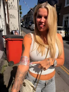 Керри Катона на улице показывает новую татуировку на предплечье. На фото видна девушка с длинными светлыми волосами, в белой топе и джинсах, указывающая пальцем на свежую татуировку на левой руке.