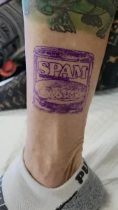 Татуировка банки SPAM на голени с зеленым принтом выше и серой ноской