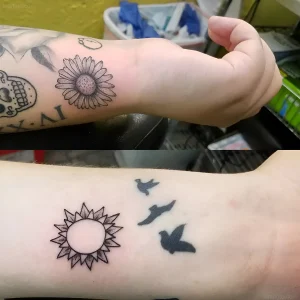 Татуировка в виде геометрического солнца и силуэтов птиц на предплечье руки, выполненная в черном цвете.