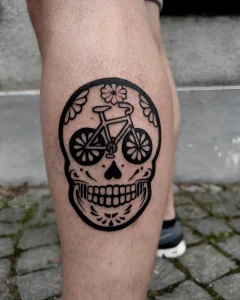 Черно-белая татуировка в стиле Sugar Skull на голени, изображающая велосипед внутри черепа с цветочными узорами