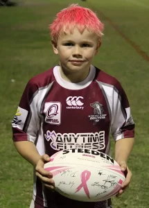 Ребёнок с ярко-розовыми волосами в игровой форме команды Canterbury Bulldogs держит регбийный мяч с розовой лентой и автографами, на фоне зелёного поля — метафора надежды и памяти о борьбе с раком