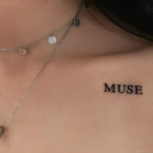 Крупный план женской груди с татуировкой слова MUSE в шрифте с засечками и тонкими цепочками с круглыми подвесками