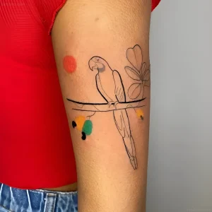 Татуировка попугая в стиле минимализм на руке девушки в красной футболке, с цветными акцентами и растительным орнаментом