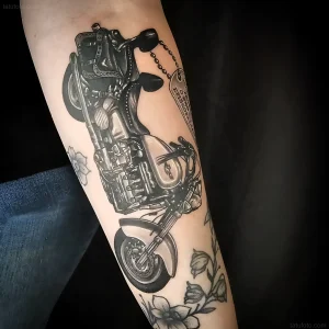Черно-белая реалистичная татуировка мотоцикла с детализированным двигателем, рулем и биркой с текстом на предплечье человека. Вокруг композиции — нежные цветы, дополняющие образ. Идеальный пример тату для байкеров ко Всемирному дню мотоциклиста.