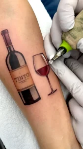 Тату-мастер в перчатках делает татуировку с надписью TINTO на бутылке вина и бокалом красного вина на предплечье клиента