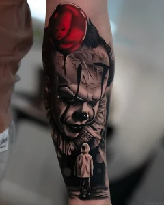 Черно-белая реалистичная татуировка Pennywise с красным шаром на предплечье, где на переднем плане стоит маленький ребенок в капюшоне, глядящий на клоуна