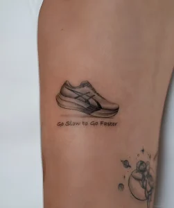 Чёрно-белая реалистичная татуировка спортивного кроссовка Asics с текстом 'Go Slow to Go Faster' на бедре человека; стиль графика, детализированная прорисовка подошвы и логотипа бренда