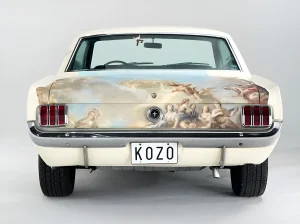 Задняя часть белого Ford Mustang 1967 года с уникальной татуировкой в стиле микро-реализм, изображающей классическую группу людей и ангелов. На бампере видна надпись 'KOZO'.