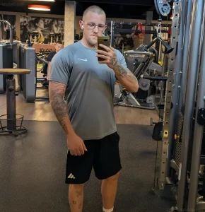 Мужчина с татуировками на руках и ногах делает селфи в зеркале спортзала, демонстрируя стильный образ для фотосессий.