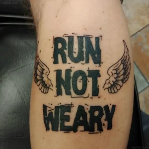 Черно-белая татуировка с надписью RUN NOT WEARY и двумя крыльями по бокам на бедре человека, выполненная в стиле old school с элементами neo-traditional
