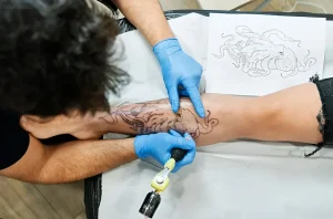 Тату-мастер в синих перчатках наносит черную татуировку с изображением осьминога на икра клиента. На столе лежит трафарет с контуром рисунка.