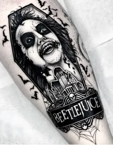 Черно-белая татуировка Битлджуса с надписью BEETLEJUICE, летучими мышами и домом Де Де на предплечье