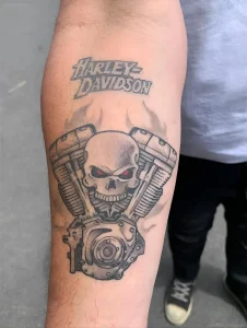 Черно-белая татуировка в стиле реализм на предплечье: мотор мотоцикла Harley-Davidson с черепом посередине и красными глазами