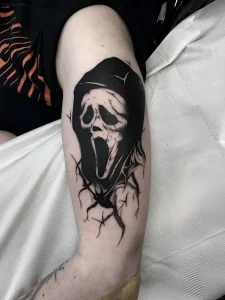 Черно-белая татуировка персонажа Ghostface из фильма «Крик» на предплечье человека, выполненная в стиле blackwork с элементами реализма и графического дизайна
