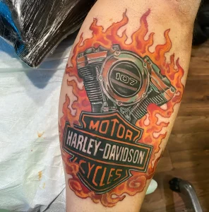 Яркая татуировка на бедре с логотипом Harley-Davidson, двигателем 107 и огненными языками пламени в стиле реализм