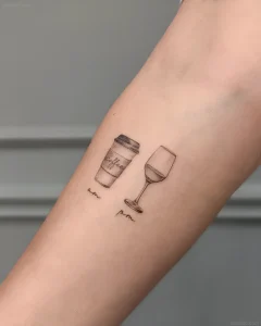 Черно-белая татуировка на предплечье: стаканчик кофе с надписью 'coffee' и бокал вина, под ними подписи 'am' и 'pm'. Минималистичный стиль, акварельная техника.