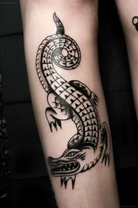 Черно-белая татуировка крокодила в стиле blackwork с геометрическим узором на предплечье, выполненная в технике плотного закрашивания и четких контуров