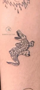 Черно-белая татуировка крокодила в стиле лайнворк на коже, видны детали чешуи и открытая пасть, сверху частично виден другой рисунок