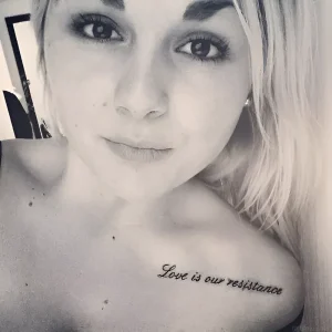 Чёрно-белое фото девушки с татуировкой 'Love is our resistance' на левой ключице, выполненной тонким курсивным шрифтом — пример тату для Дня муз 17 июня