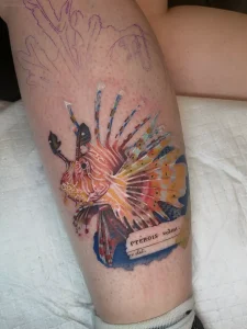 Яркая татуировка морского льва (Pterois volans) в акварельном стиле на предплечье человека, выполненная с детализацией плавников и цветовой палитрой от оранжевого до синего, с подписью латинского названия вида
