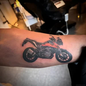 Свежая татуировка с изображением спортивного мотоцикла KTM на предплечье мужчины, выполненная в черно-белом стиле с акцентами оранжевого цвета