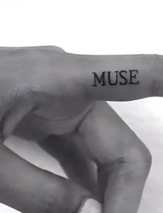 Черно-белое фото татуировки с надписью MUSE на пальце руки, выполненной в стиле минимализм