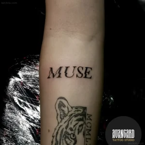 Чёрно-белая татуировка с надписью MUSE на предплечье, выполненная в стиле минимализма; ниже видна часть тигра с римскими цифрами — пример тематической татуировки к празднику 17 июня