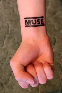 Черно-белая татуировка с надписью MUSE между двумя горизонтальными линиями на внутреннем запястье человека, выполненная в стиле stencil или lettering