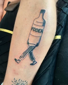 Черно-белая татуировка на руке, изображающая бутылку водки с надписью VODKA, у которой вместо туловища ноги в полосатых штанах и кроссовки