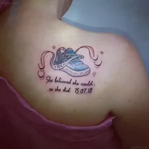 Яркая татуировка на плече в стиле нео-традишн: синяя беговая кроссовка, лента с надписью +10K, звезды и мотивирующая фраза "She believed she could... so she did" с датой 15.07.18